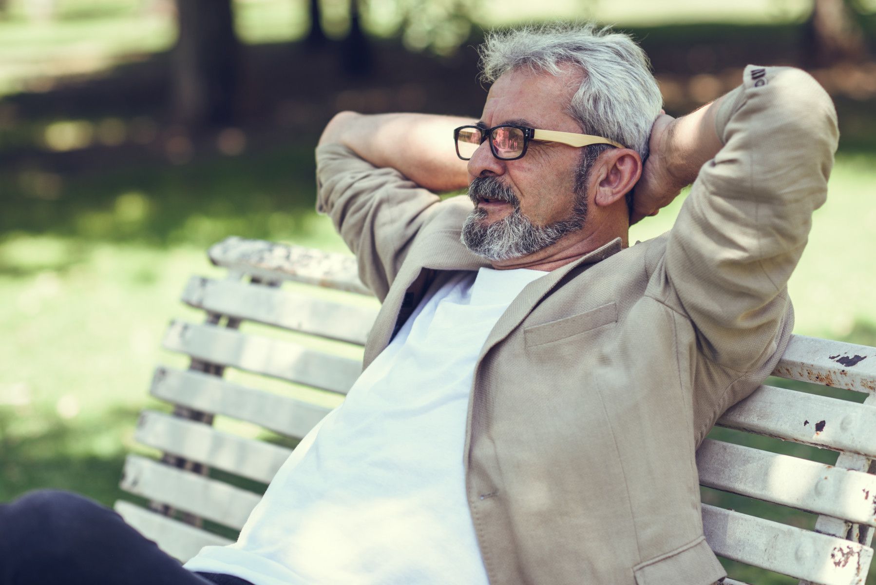 Portrait of a pensive mature man sitting on a bench in an urban park. Senior male with white hair and beard smiling wearing casual clothes. Old man wearing eyeglasses.